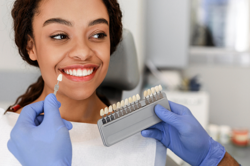 beautiful young black woman sitting in dentist chair, smiling while dentist holds a veneer up to her teeth
