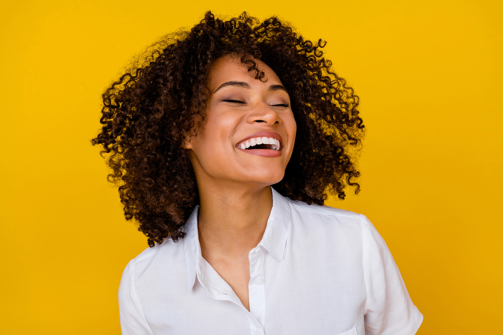 A person with curly hair is laughing joyfully, showcasing a bright smile while wearing a white shirt against a yellow background, perhaps having just enjoyed the benefits of general dentistry.