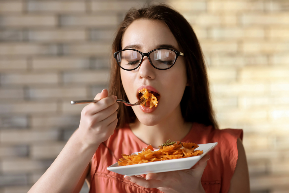 A woman with glasses eats pasta from a white plate using a fork. She is wearing a coral top and standing in front of a brick-patterned wall.