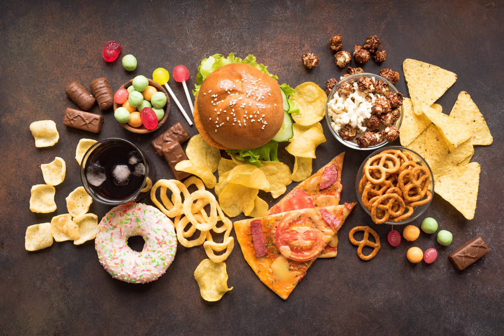 Assortment of junk food including a burger, pizza, chips, pretzels, doughnut, candy, popcorn, chocolate, and a glass of soda on a dark background.