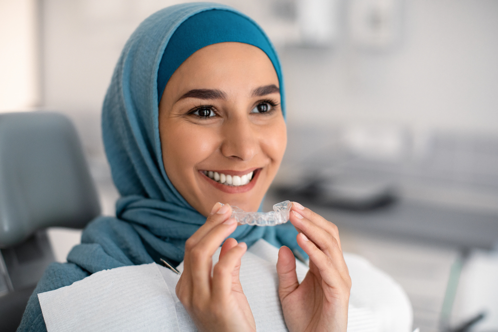 A smiling woman in a dental office holds a clear aligner near her mouth. She is wearing a light blue hijab and a dental bib.