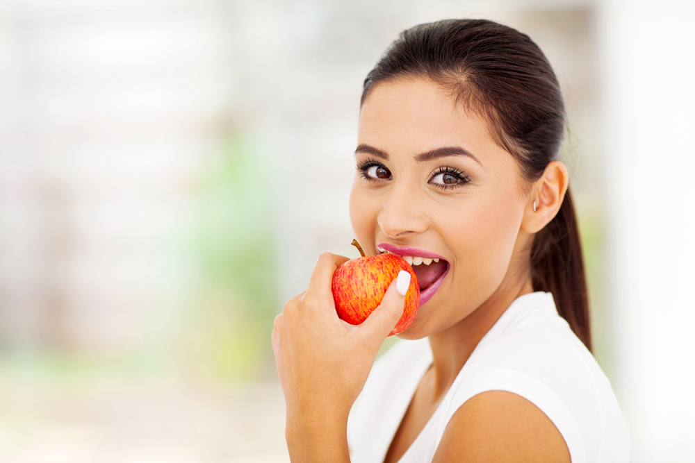A woman with dark hair in a ponytail smiles while holding a red apple near her mouth, appearing to take a bite.