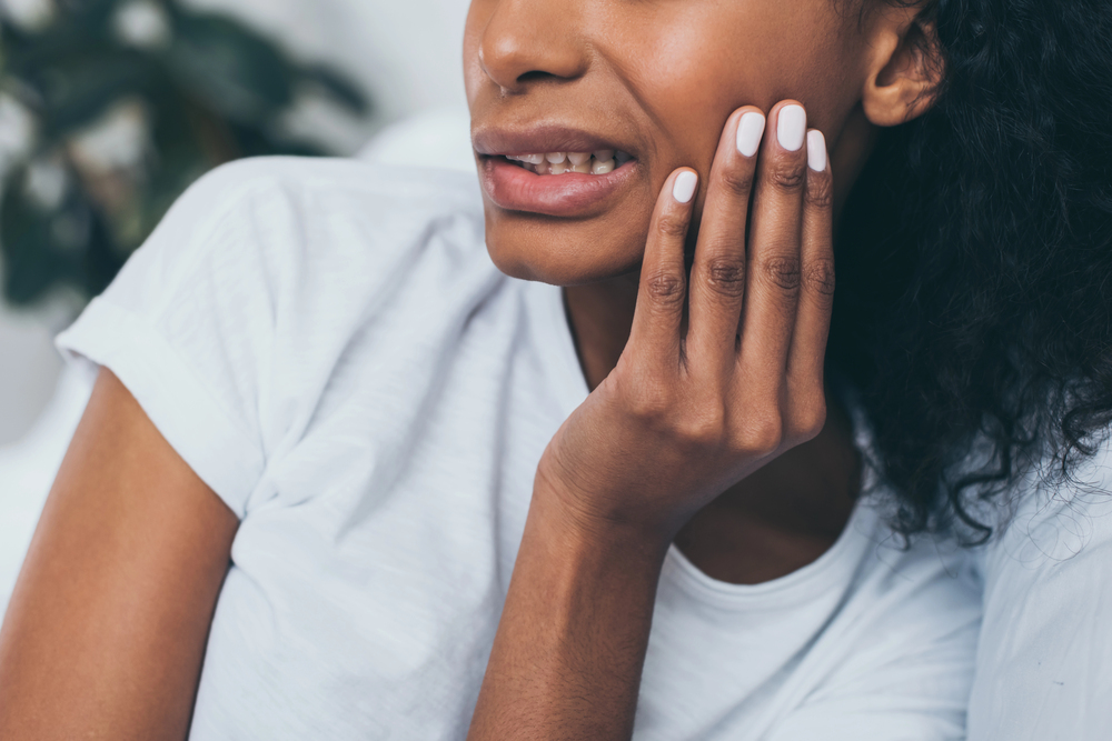 A woman in a white shirt holds her cheek and winces in pain, suggesting discomfort, possibly from a toothache or jaw pain.