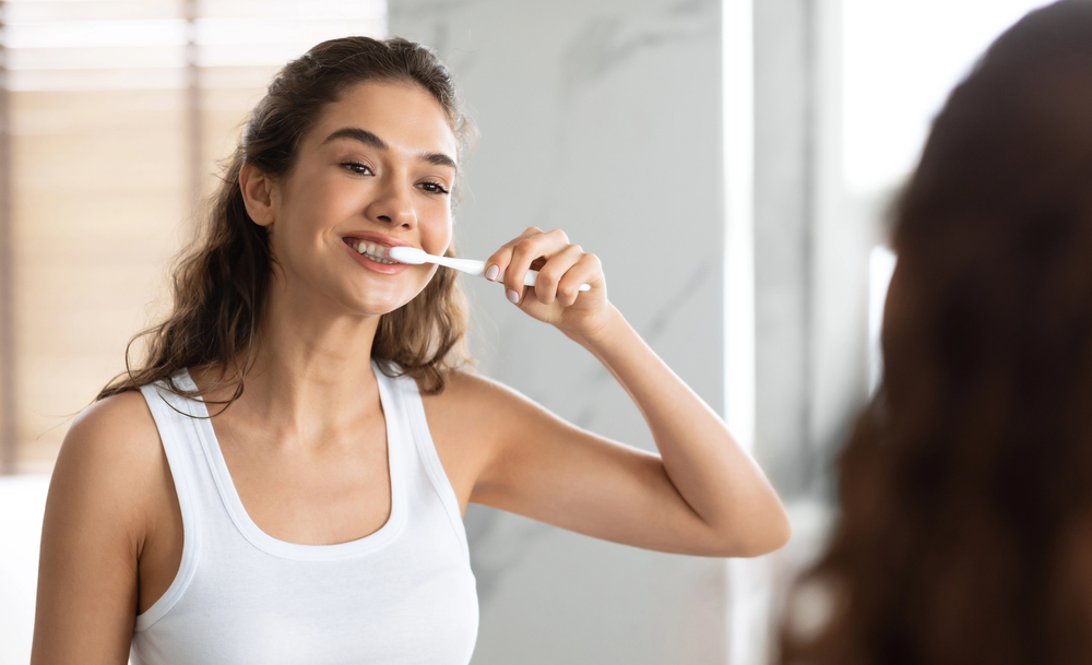 A young woman wearing a white tank top is brushing her teeth and smiling while looking in the bathroom mirror.