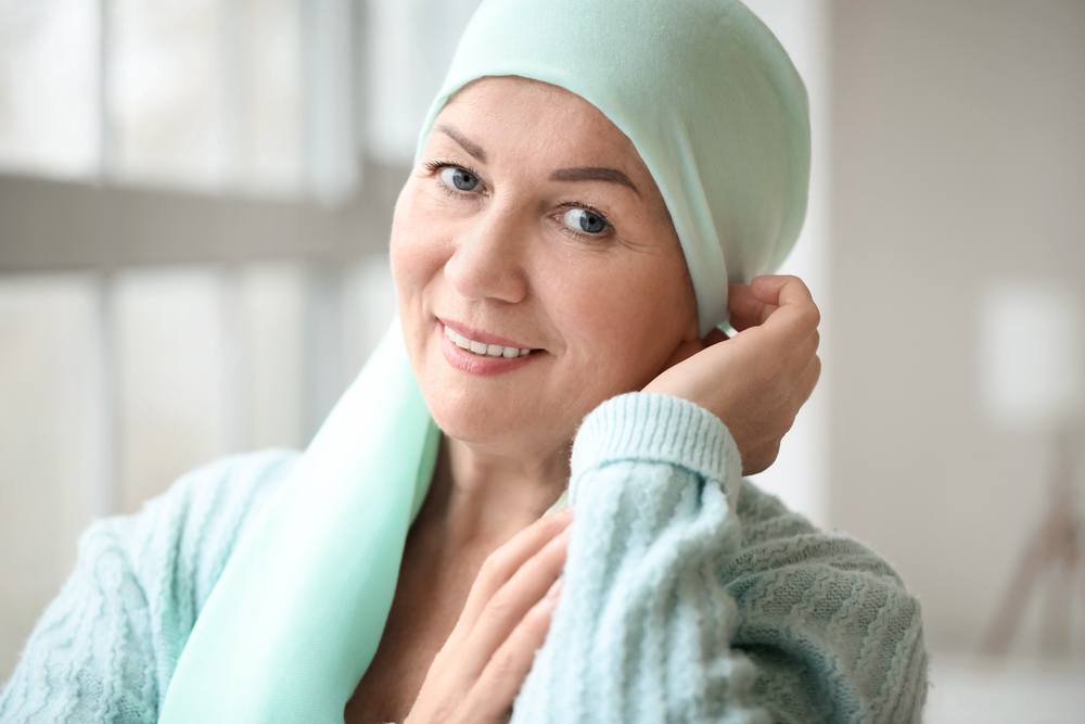 A woman wearing a light blue headscarf and sweater smiles while adjusting her headscarf, standing indoors near a window.
