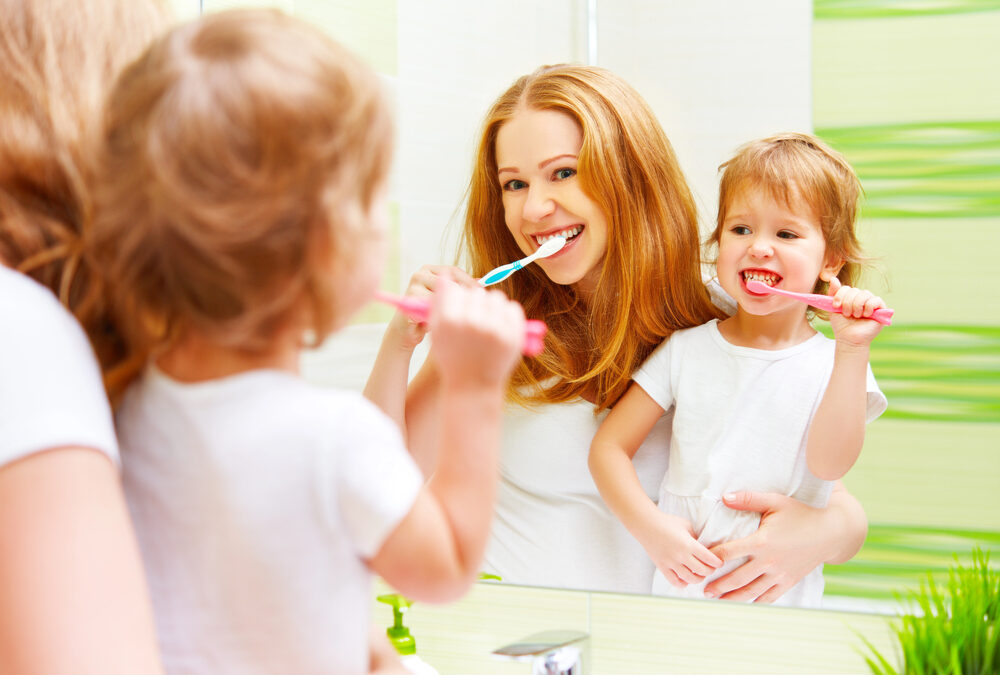 An adult and a young child brush their teeth together while standing in front of a bathroom mirror.