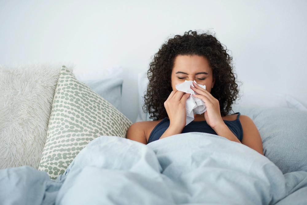 A woman sits up in bed, holding a tissue to her nose as if she is sneezing or blowing her nose. She appears to be unwell. Pillows and blankets are visible.
