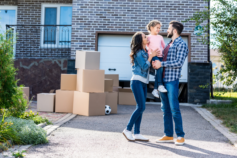 A man and woman stand in a driveway holding a child, with cardboard boxes stacked nearby in front of a house and garage.