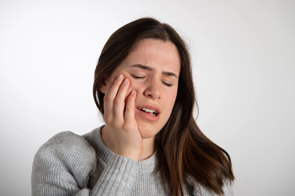 A woman with long brown hair touches her cheek and closes her eyes, appearing to be in pain, possibly from wisdom teeth or jaw discomfort.