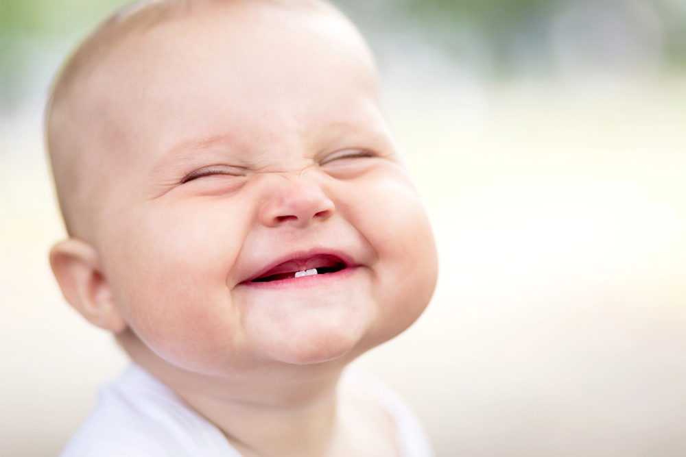 A close-up of a smiling baby with eyes closed, showing two lower teeth and wearing a white shirt. The softly blurred background highlights this milestone moment—reminding parents to ask: When should a child have their first dental visit?.