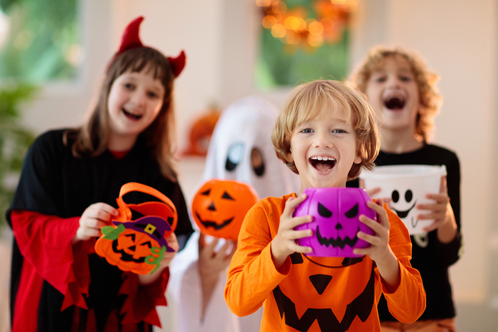 Three children in Halloween costumes hold pumpkin buckets and smile indoors; one is dressed as a devil, another as a ghost, and the third in an orange pumpkin-themed outfit.
