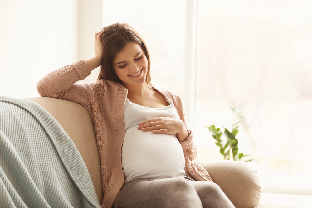 A pregnant woman sits on a sofa, smiling and resting her hand on her belly, with a soft blanket draped nearby and natural light coming through a window.