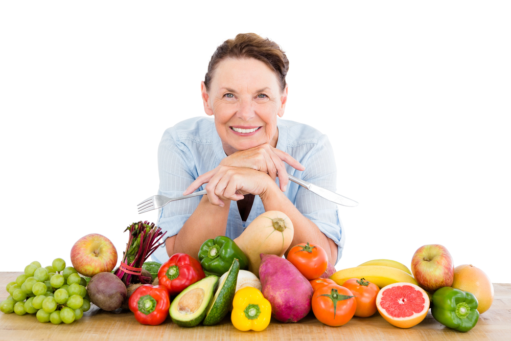 A woman with dental implants smiles behind a table filled with assorted fruits and vegetables, holding a fork and knife, with a white background.