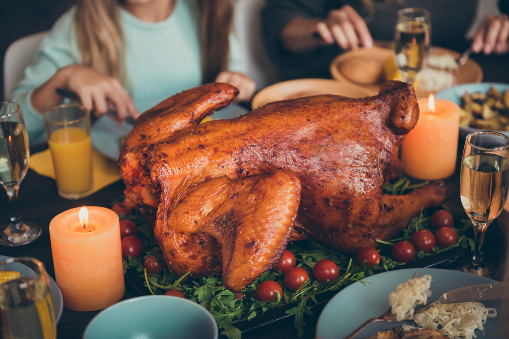 A roasted whole turkey garnished with cherry tomatoes and greens, surrounded by candles, drinks, and people at a dining table.