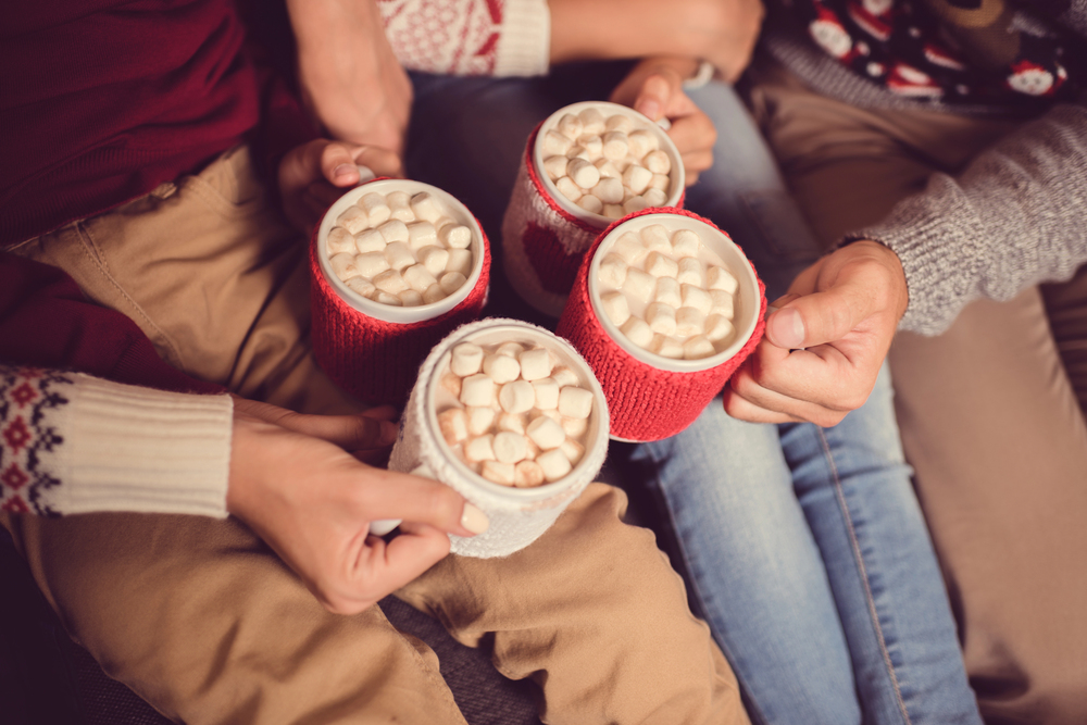 Four people holding mugs of hot chocolate topped with marshmallows, sitting closely together and wearing knitted sweaters.