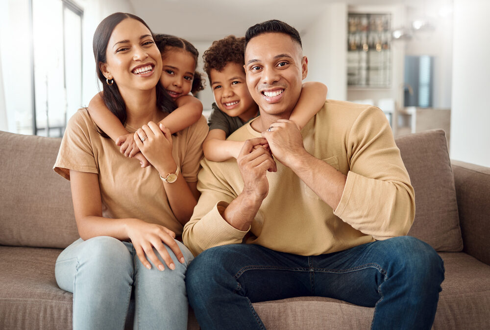 A family of four sits on a couch, smiling at the camera. Two children hug their parents from behind. The setting appears to be a modern, well-lit living room.