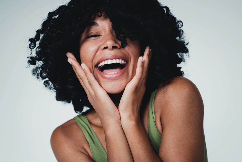 A person with curly hair smiles widely with hands on their cheeks, wearing a green sleeveless top against a plain background.
