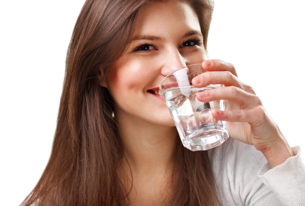 A woman with long brown hair holds a glass of water and smiles, wearing a white top against a plain white background.