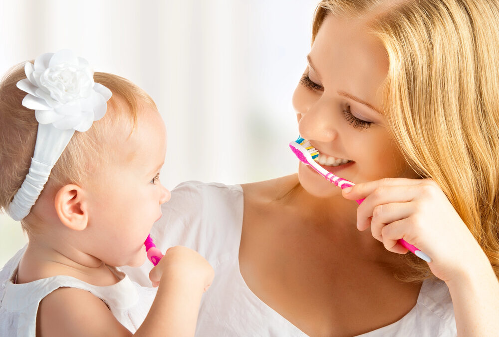 A woman and a baby, both dressed in white, are smiling and holding toothbrushes, appearing to brush their teeth together.