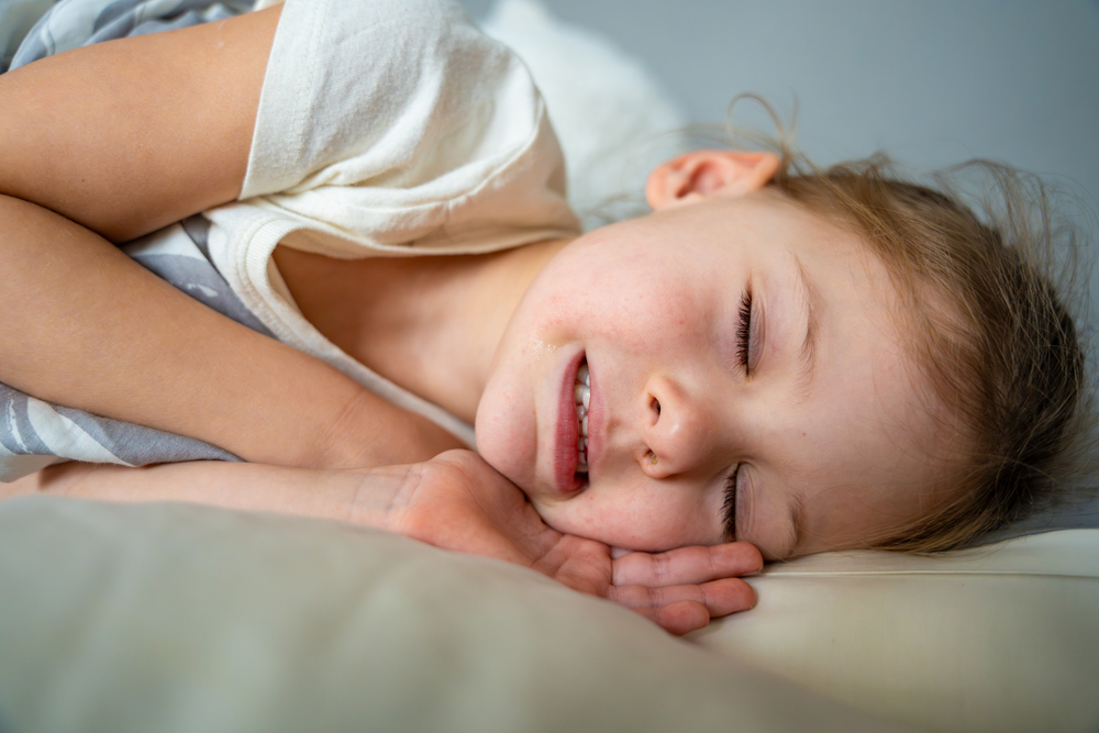 A young child with light brown hair is lying on a pillow, sleeping on their side with a relaxed expression and eyes closed.