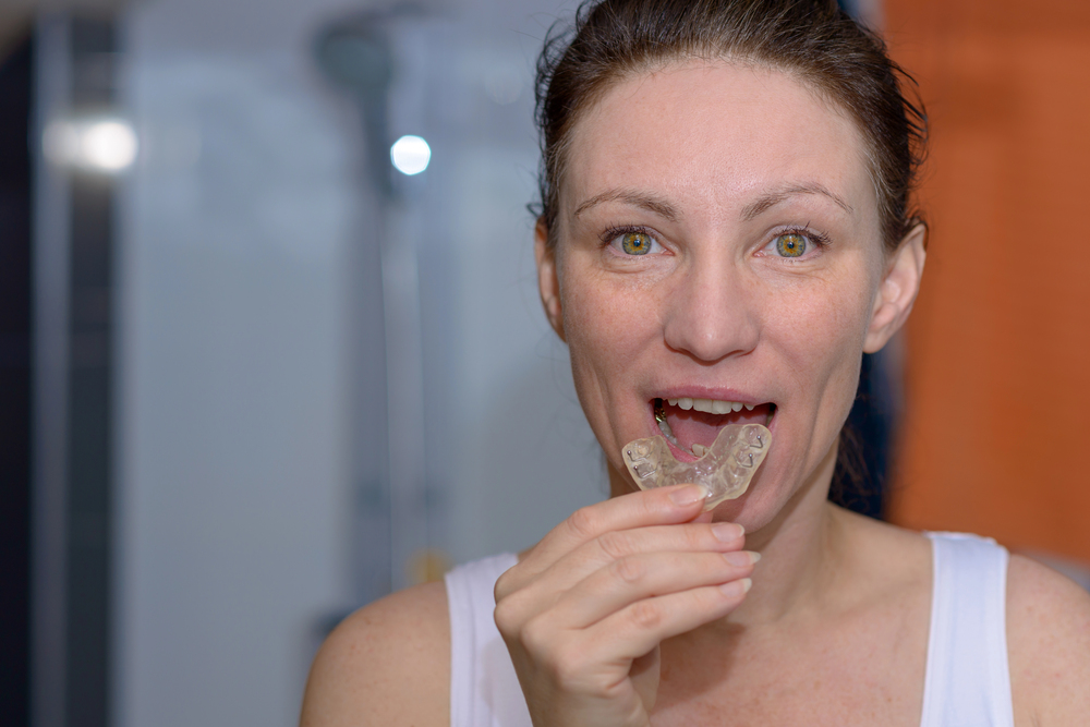 A woman with light skin is holding a clear dental mouthguard near her mouth in a bathroom, preparing to insert it.