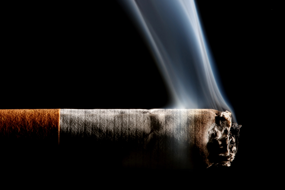 Close-up of a lit cigarette burning with smoke rising against a black background.