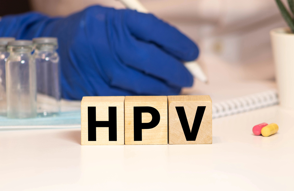 Wooden blocks spell "HPV" on a table with a gloved hand writing, medicine bottles, pills, and a notepad in the background.