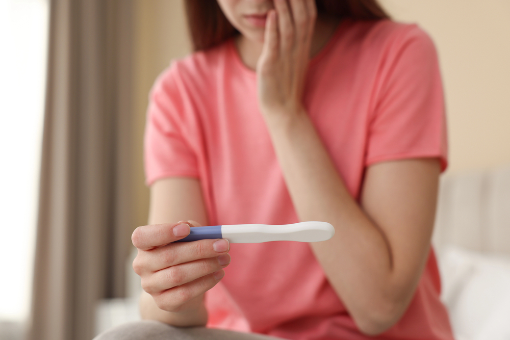 A woman in a pink shirt sits on a bed, holding a pregnancy test and touching her face with a concerned expression.