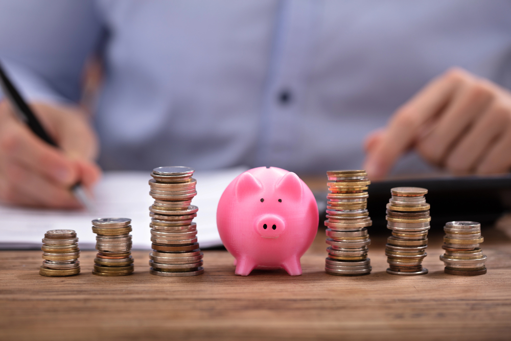 Pink piggy bank surrounded by stacked coins of varying heights, with a person in the background writing on paper and using a calculator.