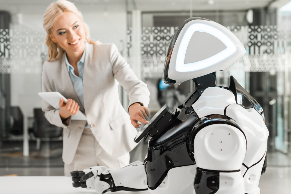 A woman in business attire interacts with a humanoid robot in an office setting while holding a tablet.