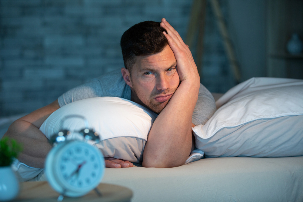 Man lying in bed with his head on a pillow, looking tired and resting his hand on his face, with an alarm clock in the foreground.