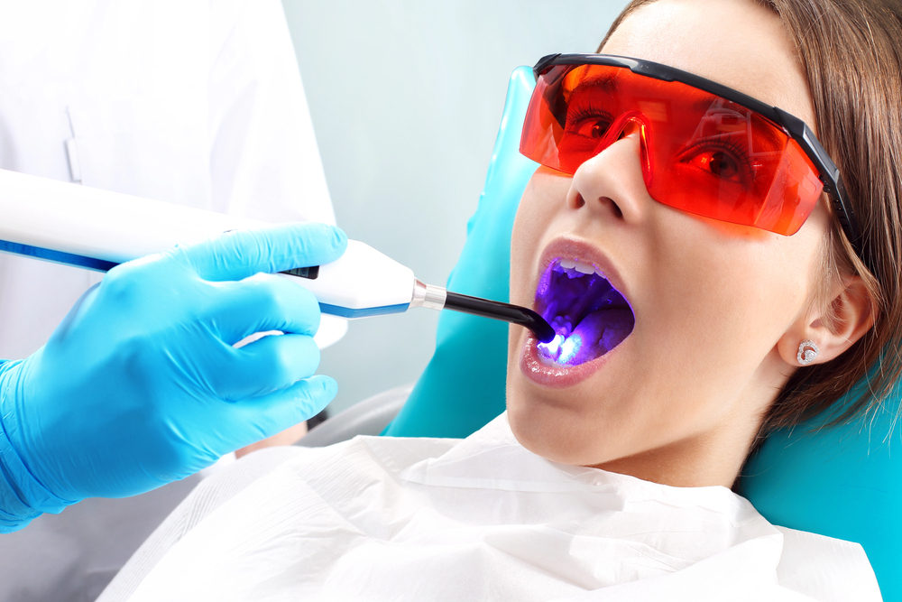 A dentist uses a curing light tool inside a patient’s mouth. The patient is wearing protective orange glasses and a dental bib.